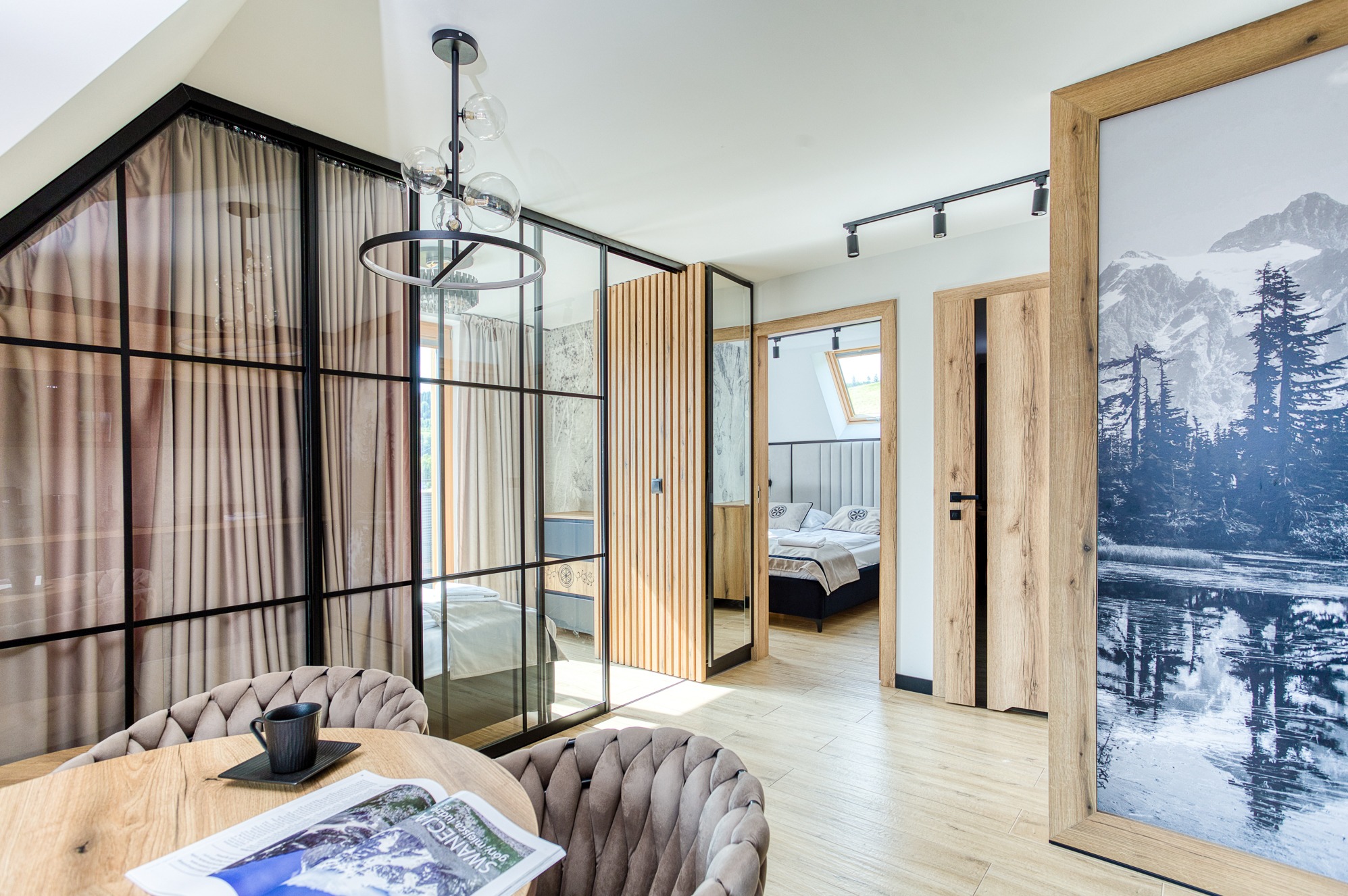 Wnętrze apartamentu z drewnianą podłogą, szklaną ścianą z zasłonami i widokiem na sypialnię. Na pierwszym planie stół z gazetą i filiżanką.