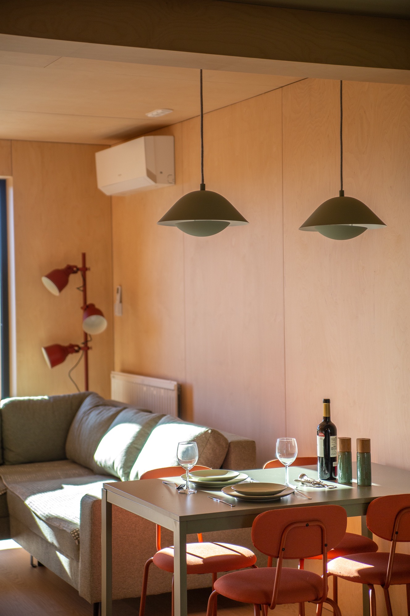 Jasne wnętrze z drewnianymi panelami: stół z nakryciem, butelka wina, sofa i lampy. Minimalistyczny design z akcentami koloru pomarańczowego.