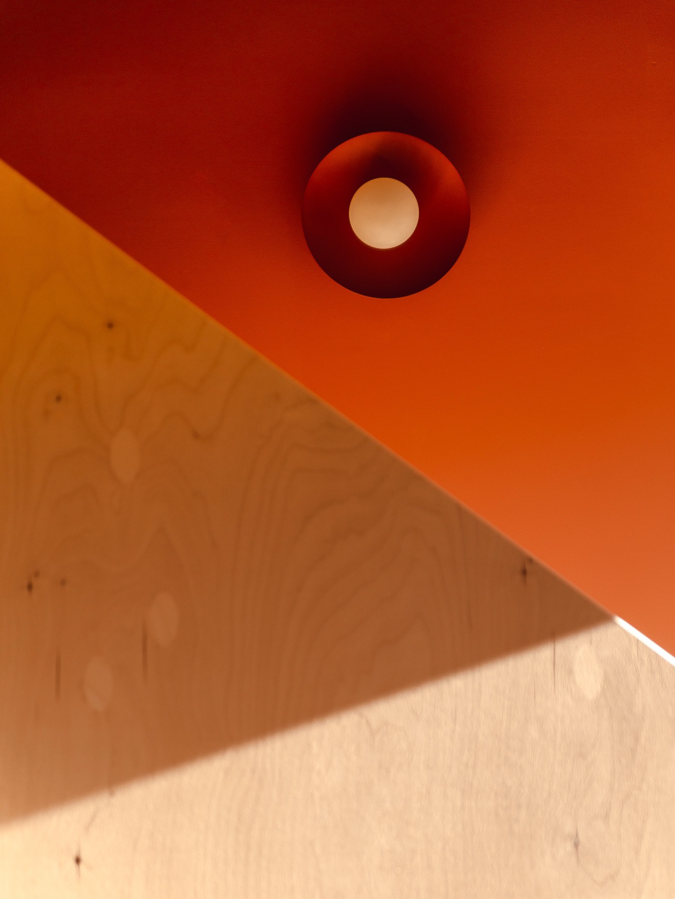 Pomarańczowy sufit z okrągłą lampą i fragmentem drewnianej ściany w ciepłym oświetleniu. Nietypowe ujęcie z dołu podkreśla geometrię wnętrza.
