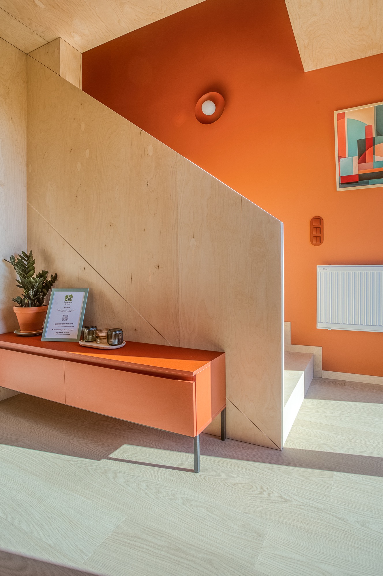 Nowoczesne wnętrze z pomarańczową ścianą, drewnianymi schodami i szafką. Na szafce roślina, ramka z informacją i dekoracyjne świeczniki. Minimalistyczny design.