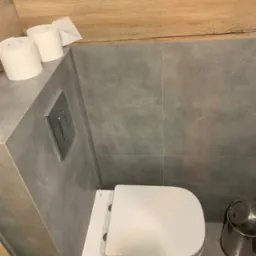 Nowoczesna toaleta wisząca z szarą armaturą i drewnianym akcentem. Minimalistyczny design łazienki z rolkami papieru toaletowego i metalowym koszem.