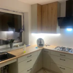 Jasna kuchnia z drewnianymi akcentami: szafki górne z fornirem, beżowe fronty dolne z czarnymi uchwytami, płyta gazowa i okno z rośliną w doniczce.