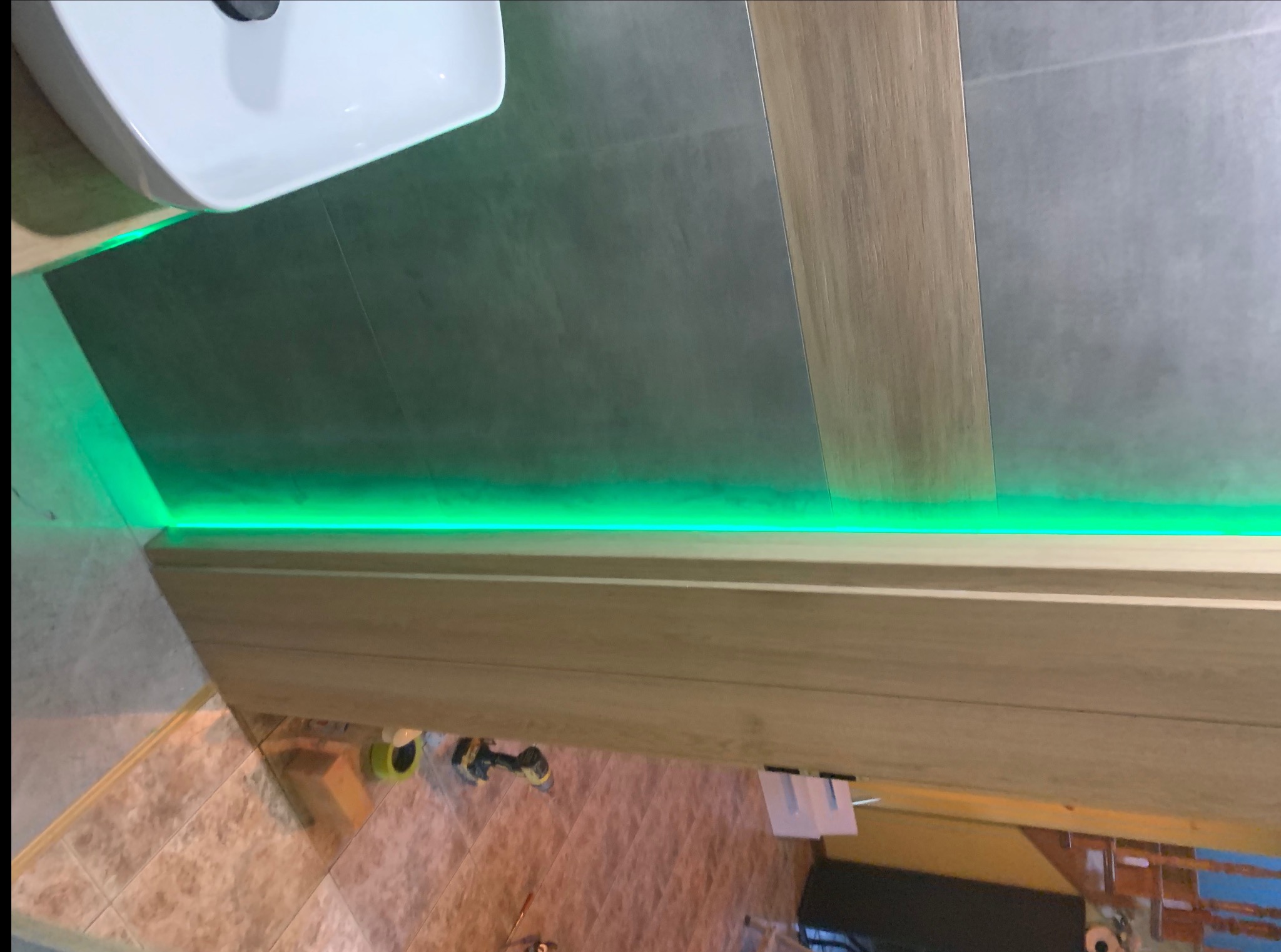 Nowoczesna łazienka z podświetlanym na zielono blatem pod umywalką, szare płytki podłogowe imitujące beton, drewnopodobne wykończenia i biała umywalka.