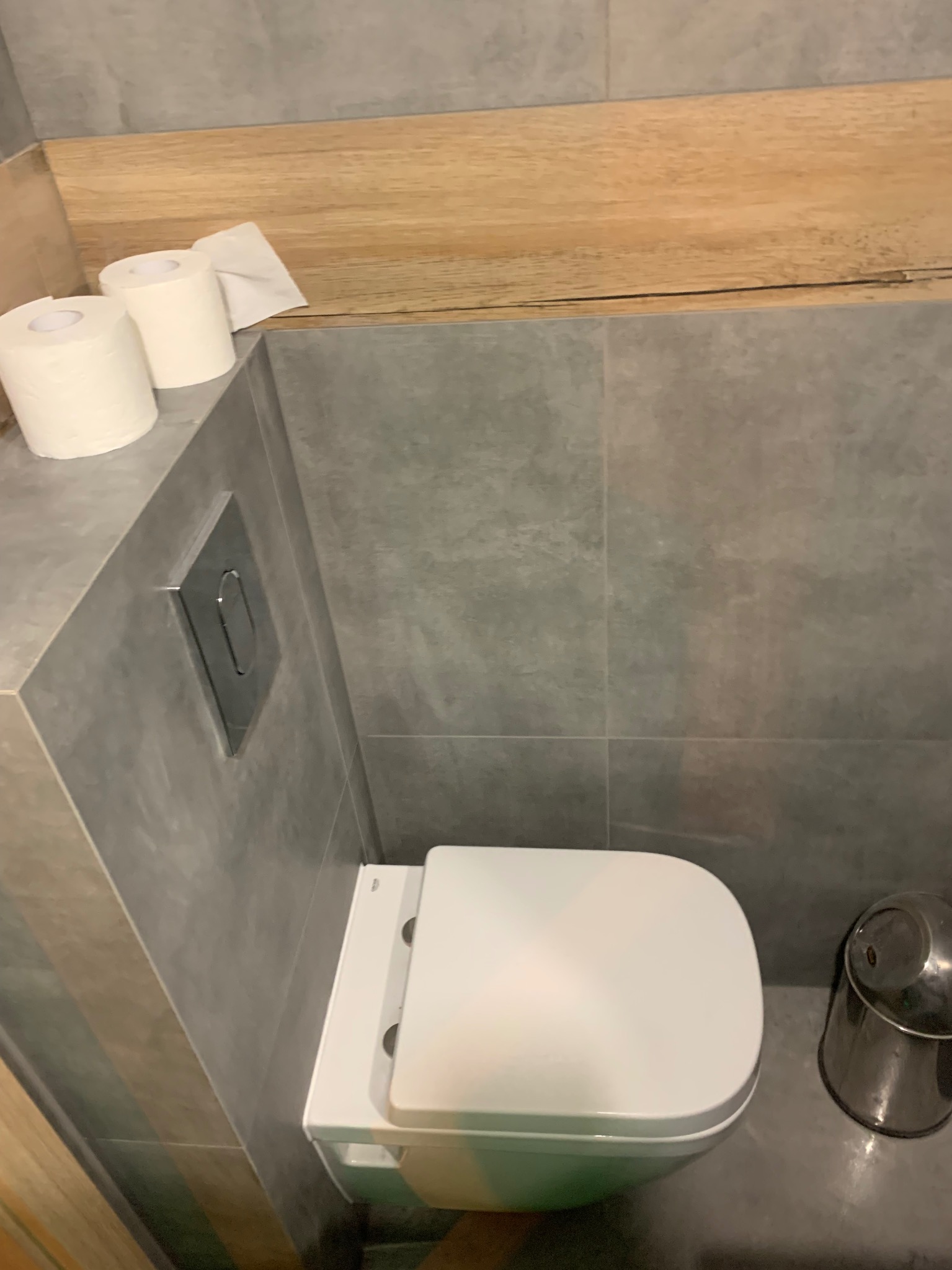 Nowoczesna toaleta wisząca z szarą armaturą i drewnianym akcentem. Minimalistyczny design łazienki z rolkami papieru toaletowego i metalowym koszem.