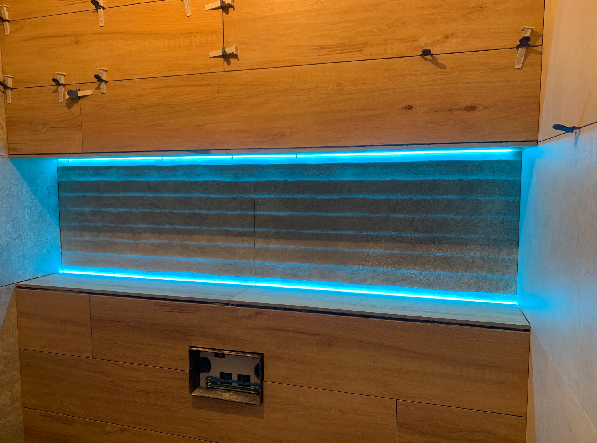 Łazienka w trakcie wykańczania: drewnopodobne płytki z niebieskim podświetleniem LED, widoczne krzyżyki glazurnicze, otwór na stelaż podtynkowy WC.