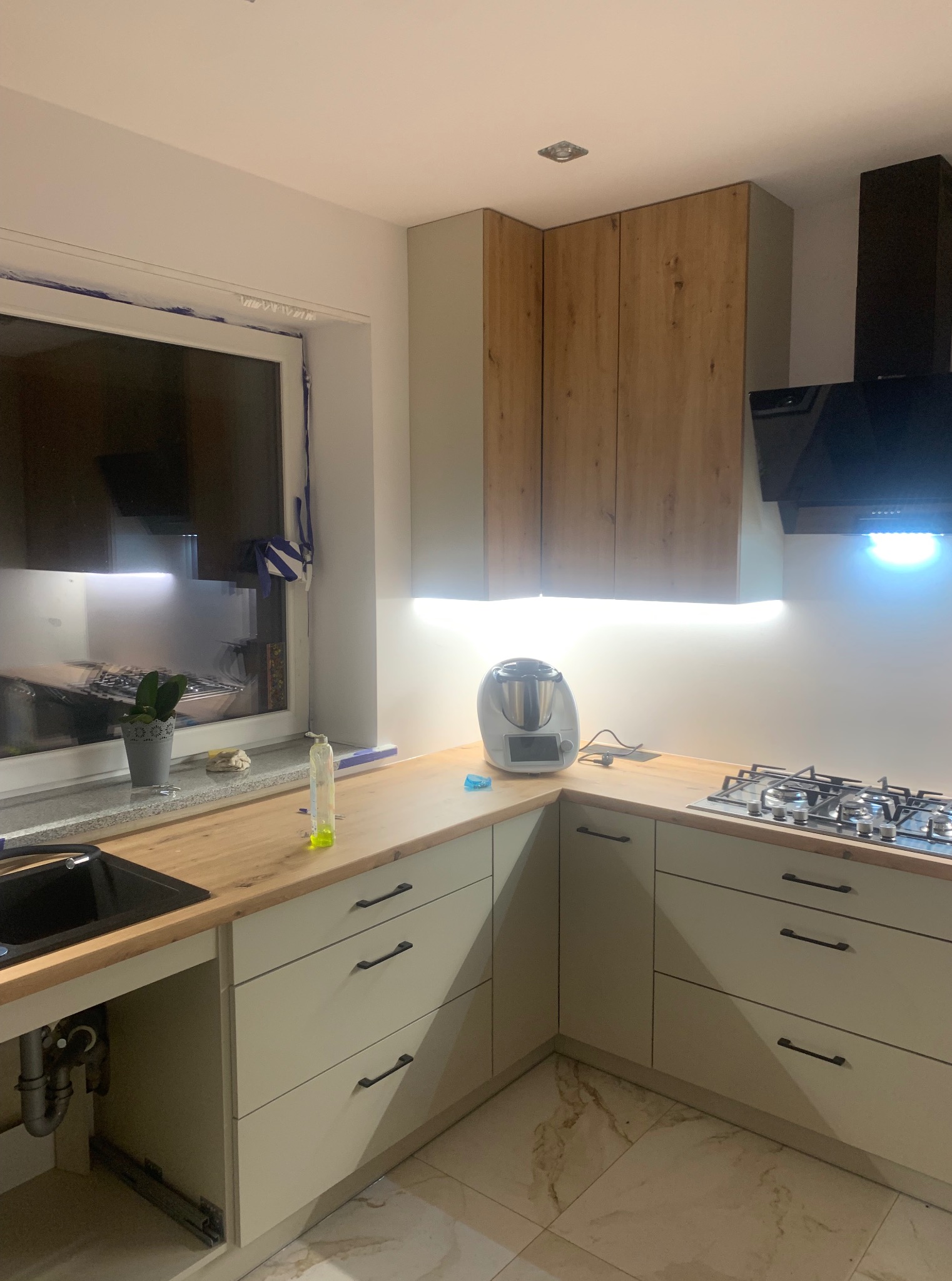 Jasna kuchnia z drewnianymi akcentami: szafki górne z fornirem, beżowe fronty dolne z czarnymi uchwytami, płyta gazowa i okno z rośliną w doniczce.