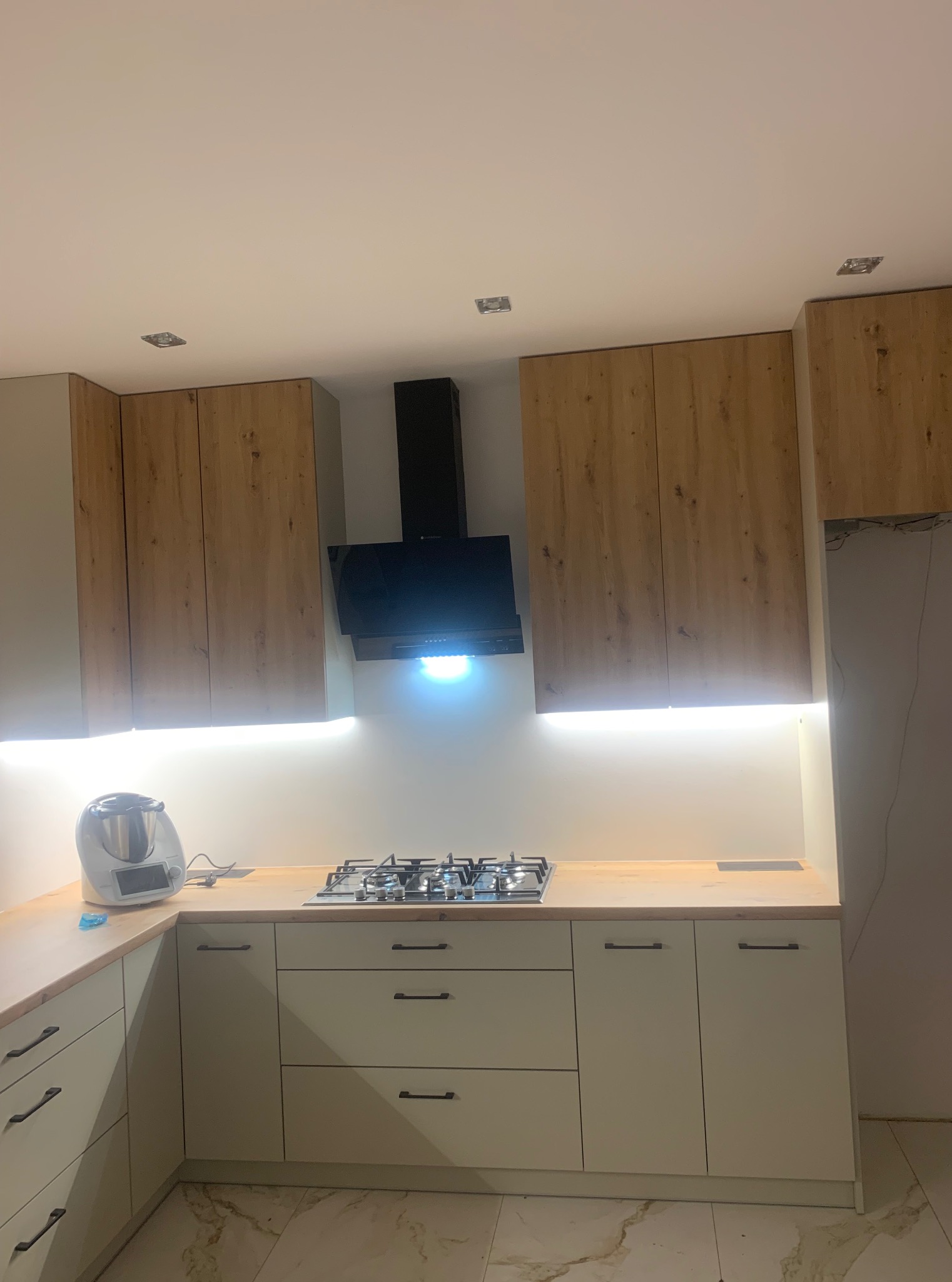 Jasna kuchnia z drewnianymi szafkami górnymi i blatem. Oświetlenie LED pod szafkami. Fragment ściany bez wykończenia po prawej stronie kadru.