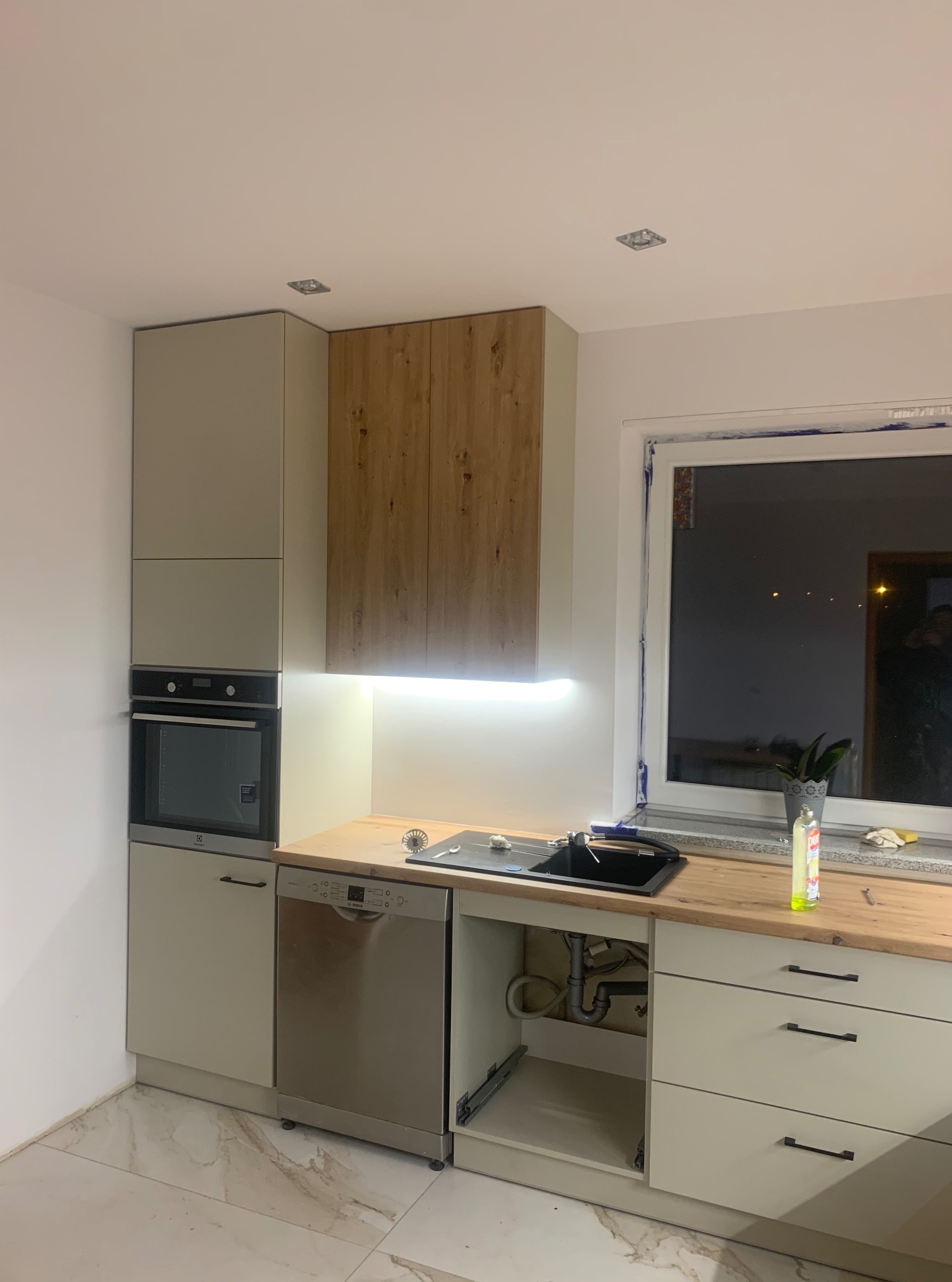 Kuchnia w Panki po remoncie: kremowe szafki z drewnianymi akcentami, wbudowany piekarnik i zmywarka, drewniany blat z czarnym zlewem, okno z rośliną.