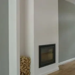 Biała zabudowa kominka z wnęką na drewno i czarnym wkładem kominkowym. Podłoga z paneli. Ściany w odcieniach bieli i szarości. Minimalistyczny design.