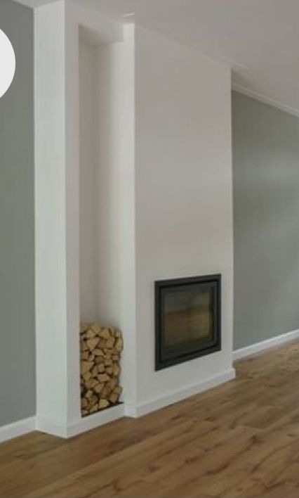 Biała zabudowa kominka z wnęką na drewno i czarnym wkładem kominkowym. Podłoga z paneli. Ściany w odcieniach bieli i szarości. Minimalistyczny design.