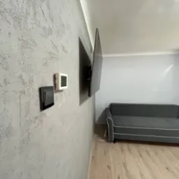 Ściana z szarą, dekoracyjną strukturą tynku w pokoju z szarą sofą i telewizorem. Widoczne włączniki. Podłoga wyłożona panelami.