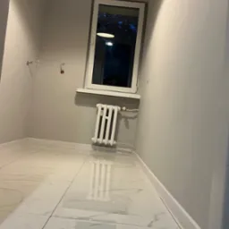 Wykończone wnętrze pokoju z marmurową podłogą i grzejnikiem pod oknem. Widoczne listwy przypodłogowe i przygotowane wyprowadzenia instalacji elektrycznej.