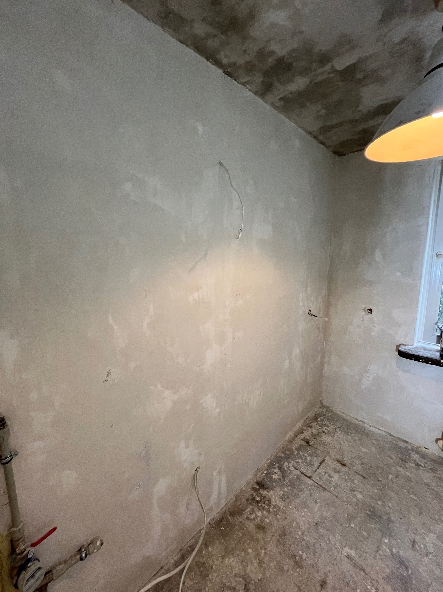 Wnętrze pokoju w trakcie remontu: ściany po szpachlowaniu, widoczne wystające kable, surowy beton na podłodze, lampa sufitowa i fragment okna z parapetem.