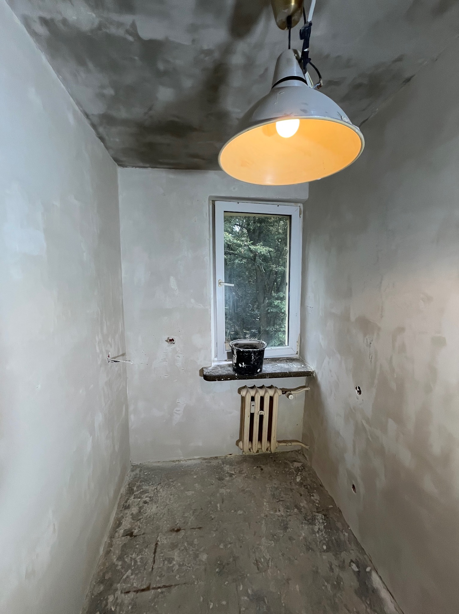 Pokój w trakcie szpachlowania: ściany pokryte warstwą gładzi, surowy beton na podłodze, wiadro na parapecie, widok z okna na zieleń, lampa wisząca.