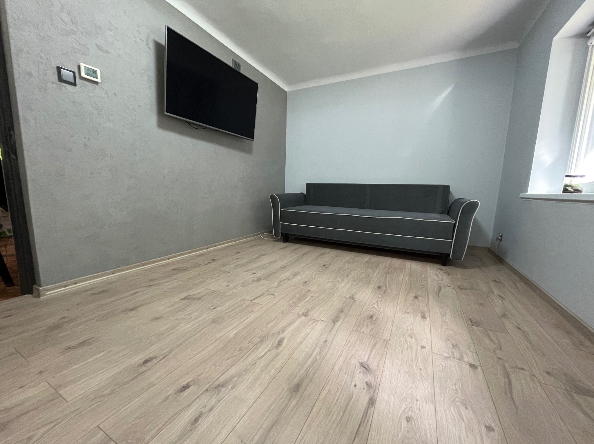 Szary salon po remoncie: ściana z efektem betonu, podłoga z paneli w odcieniach beżu, szara sofa z białymi lamówkami i wiszący telewizor.