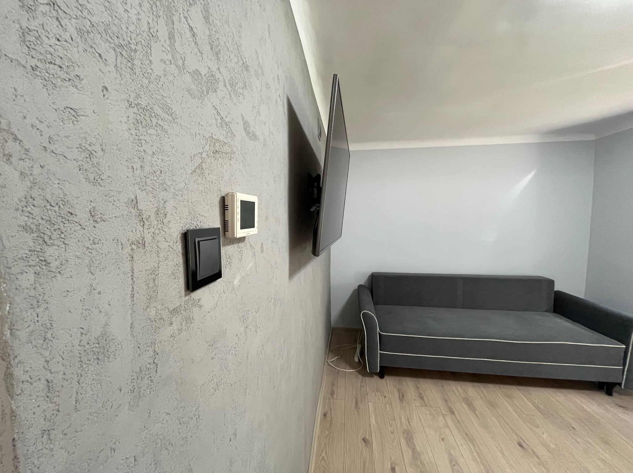 Ściana z szarą, dekoracyjną strukturą tynku w pokoju z szarą sofą i telewizorem. Widoczne włączniki. Podłoga wyłożona panelami.