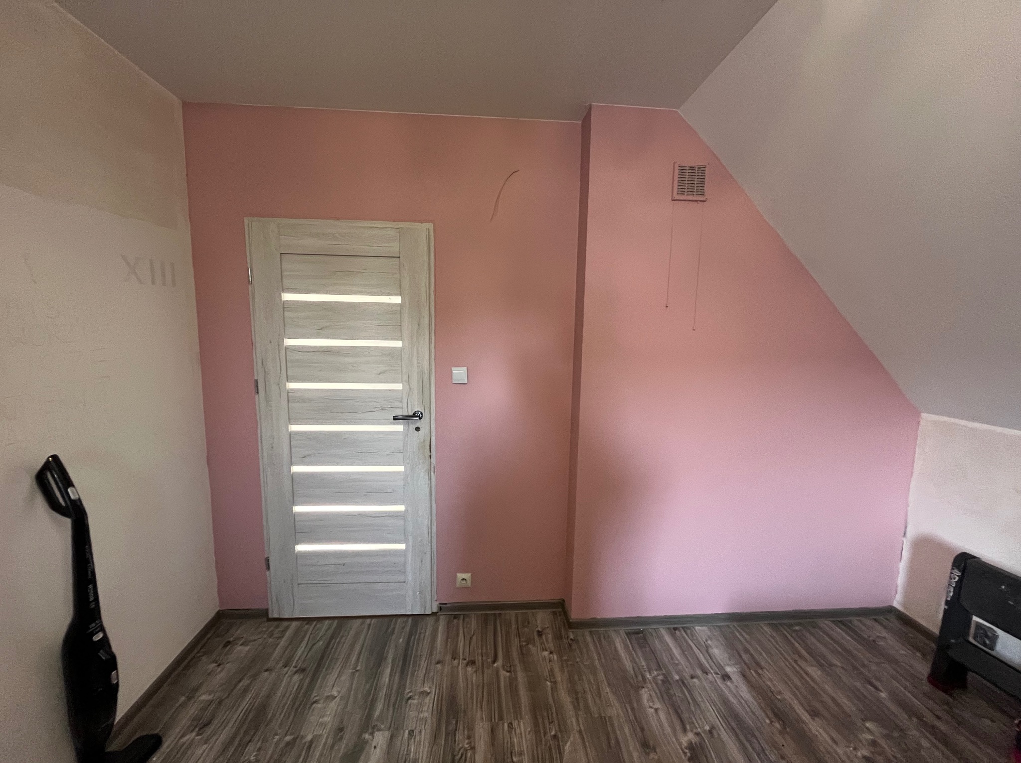 Pokój na poddaszu po malowaniu ścian na różowo, z nową podłogą imitującą drewno i białymi drzwiami z poziomymi panelami. Ściana częściowo przygotowana do malowania.