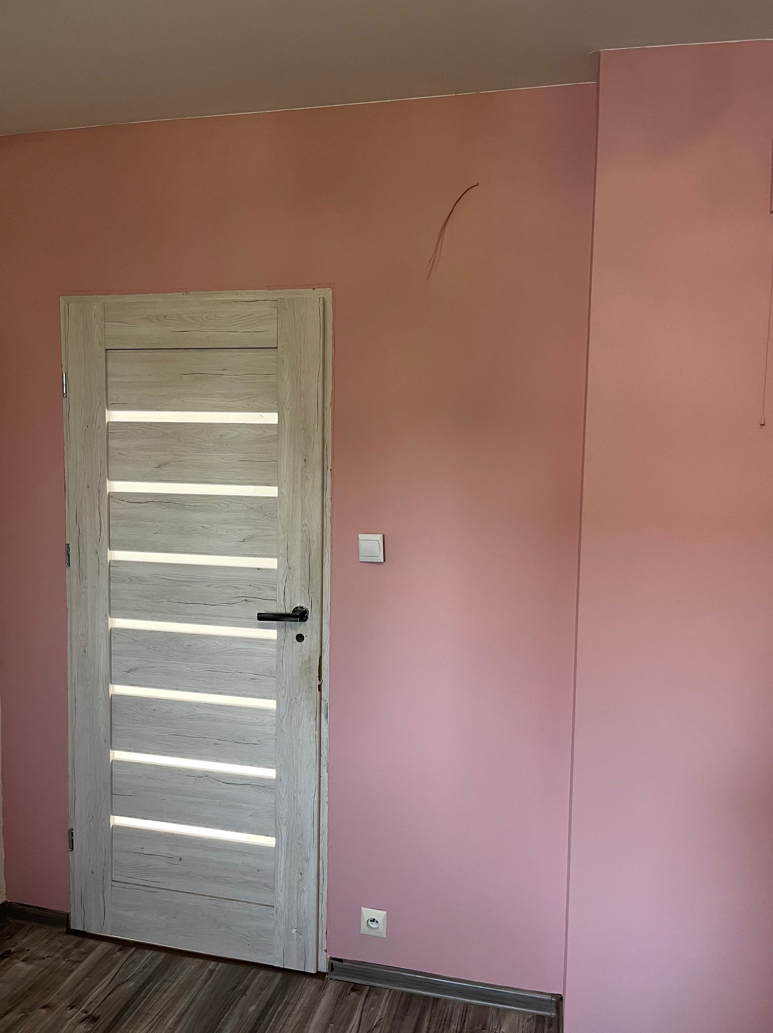 Różowa ściana po malowaniu z zamontowanymi, nowoczesnymi drzwiami z przeszkleniami. Widoczny fragment podłogi.