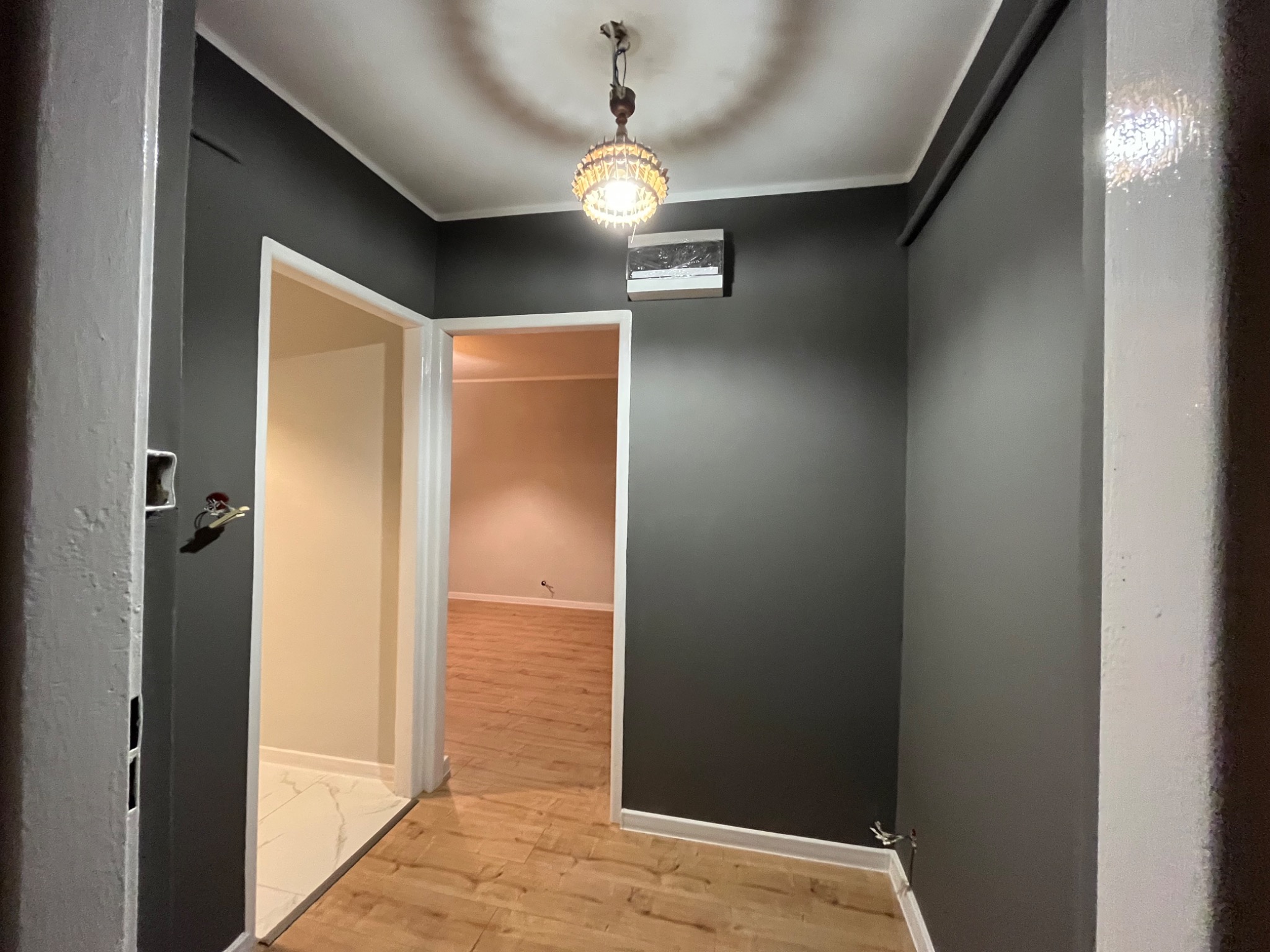 Wnętrze po remoncie: ciemnoszare ściany, drewnopodobne panele podłogowe, białe futryny drzwi i ozdobna lampa sufitowa. Widok z korytarza do pomieszczenia z jasnymi ścianami i marmurową podłogą.