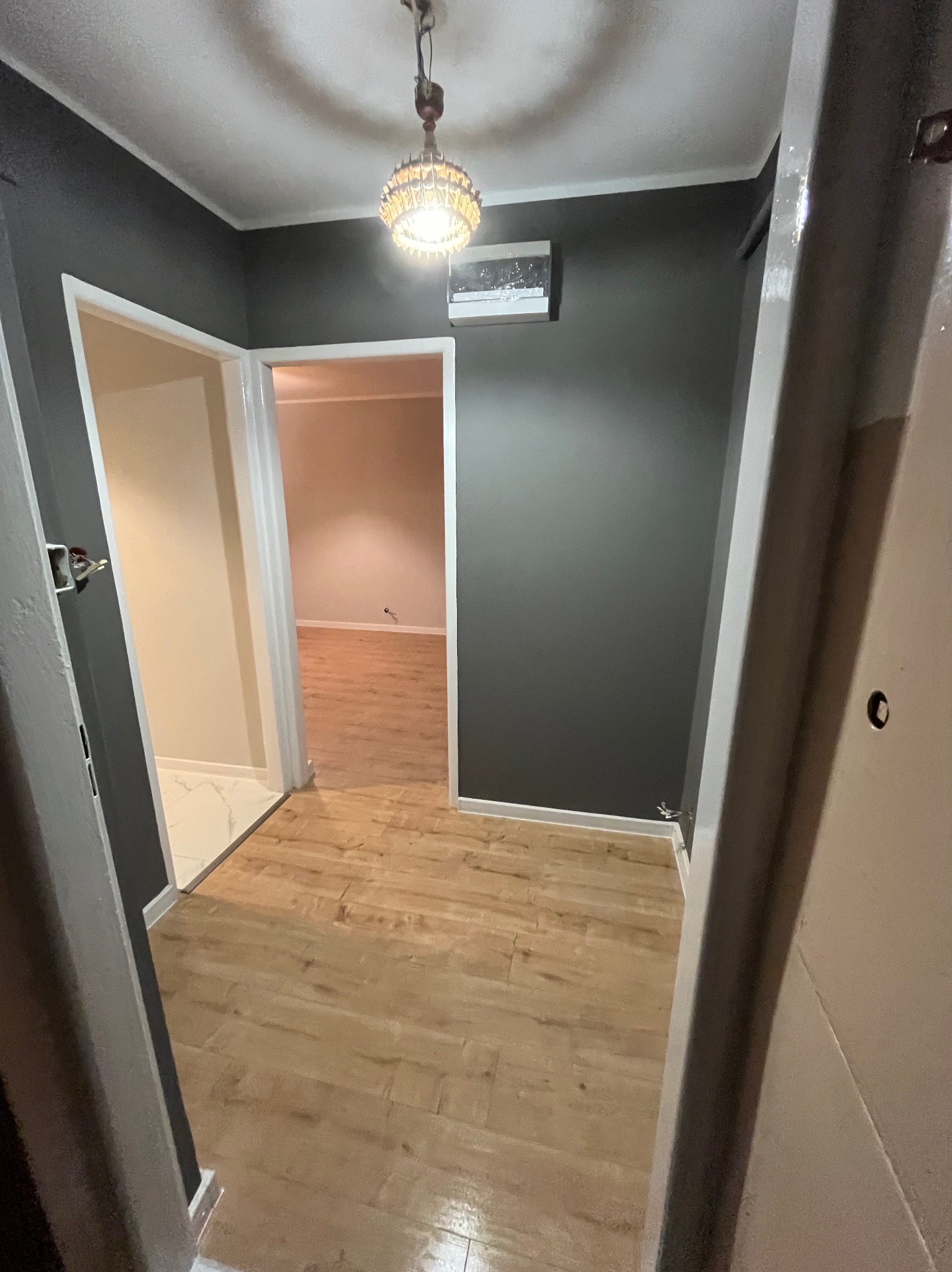 Wnętrze po remoncie: ciemnoszare ściany, panele podłogowe imitujące drewno, widok na sąsiednie pomieszczenia z białymi ścianami i marmurową podłogą. Lampa sufitowa w stylu retro.