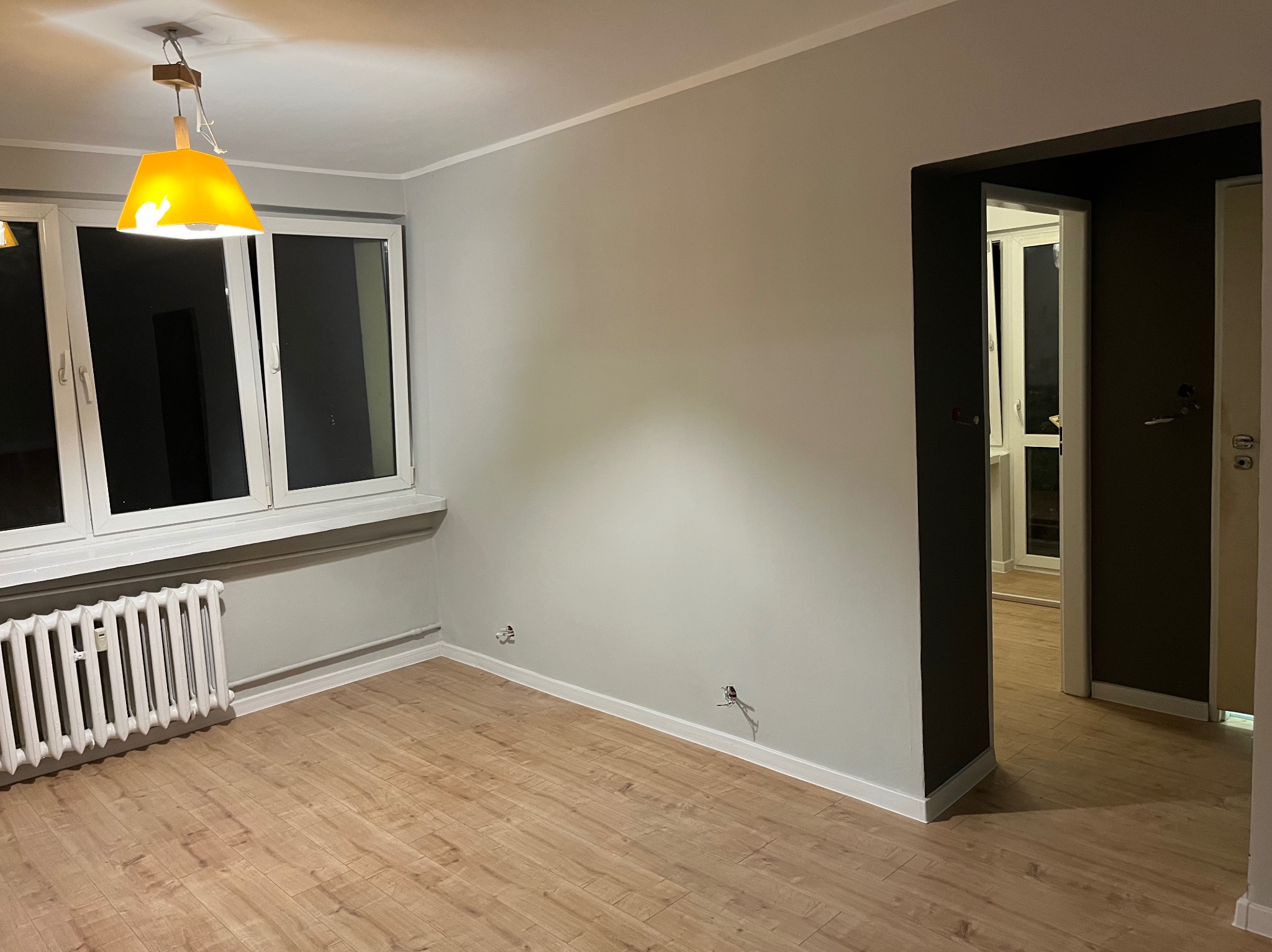 Jasne panele podłogowe, szare ściany i białe okna w pustym pokoju. Widoczny grzejnik i żółta lampa. Gniazdka elektryczne wystają ze ścian.