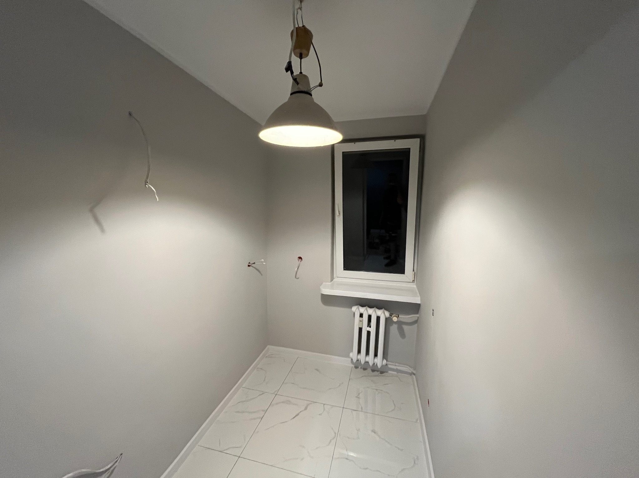 Wnętrze pomalowanego na szaro pokoju z białymi płytkami podłogowymi i wiszącą lampą. Widoczne okno, grzejnik i wystające przewody elektryczne ze ściany.