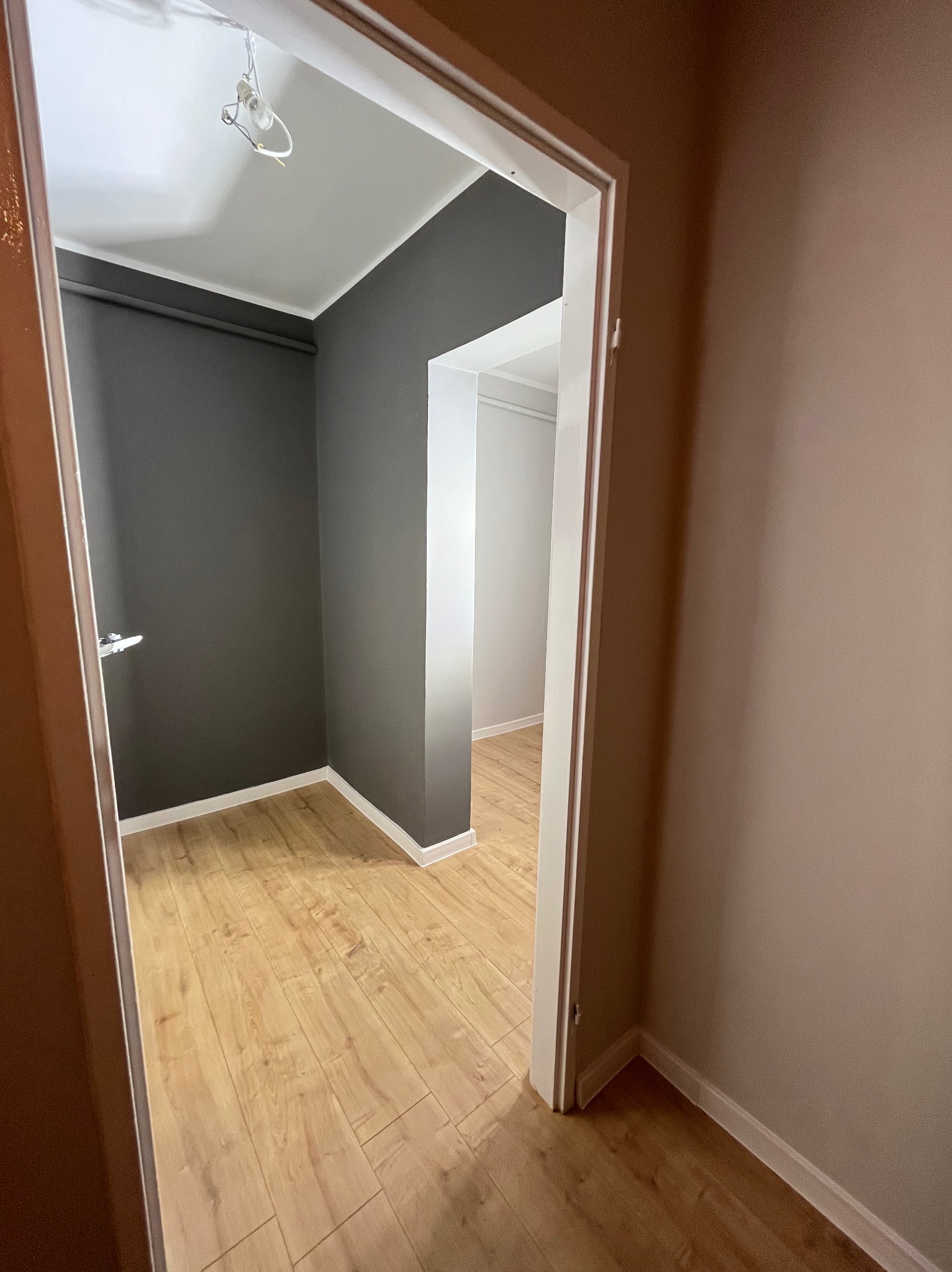 Wnętrze po remoncie: szare ściany, jasne panele podłogowe, widok przez białe futryny drzwi. Częściowo widoczna żarówka i brązowa ściana w korytarzu.