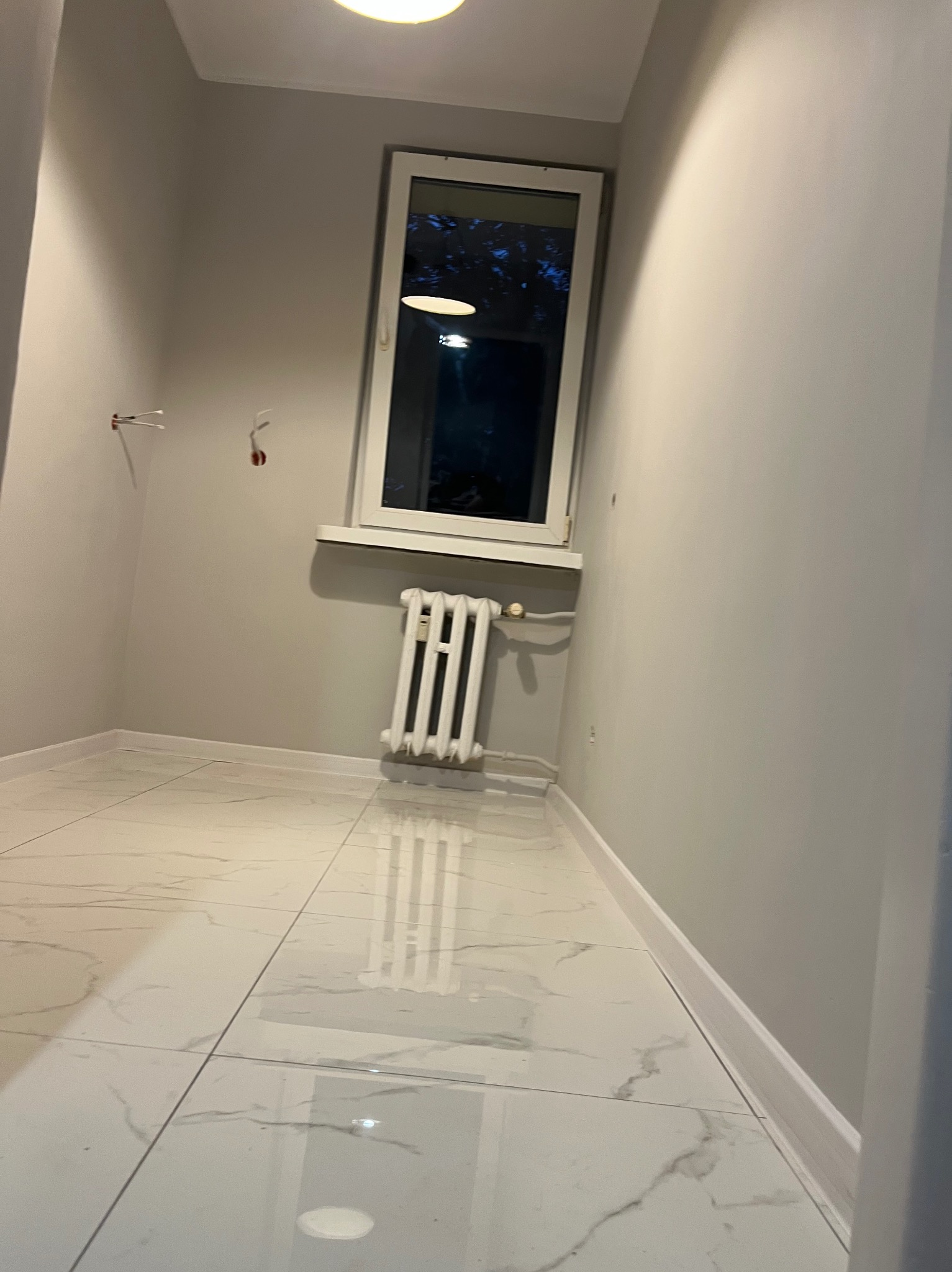 Wykończone wnętrze pokoju z marmurową podłogą i grzejnikiem pod oknem. Widoczne listwy przypodłogowe i przygotowane wyprowadzenia instalacji elektrycznej.