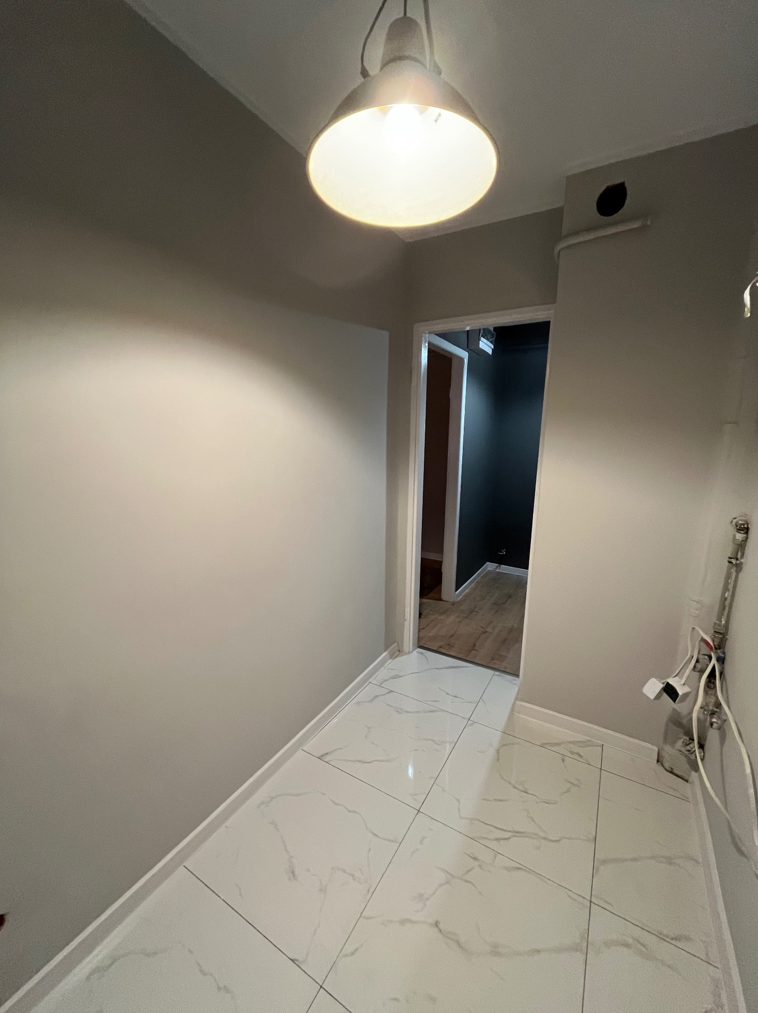 Korytarz po remoncie: gładkie, szare ściany, białe płytki podłogowe z szarym wzorem. W tle widoczne otwarte drzwi do ciemnego pomieszczenia.