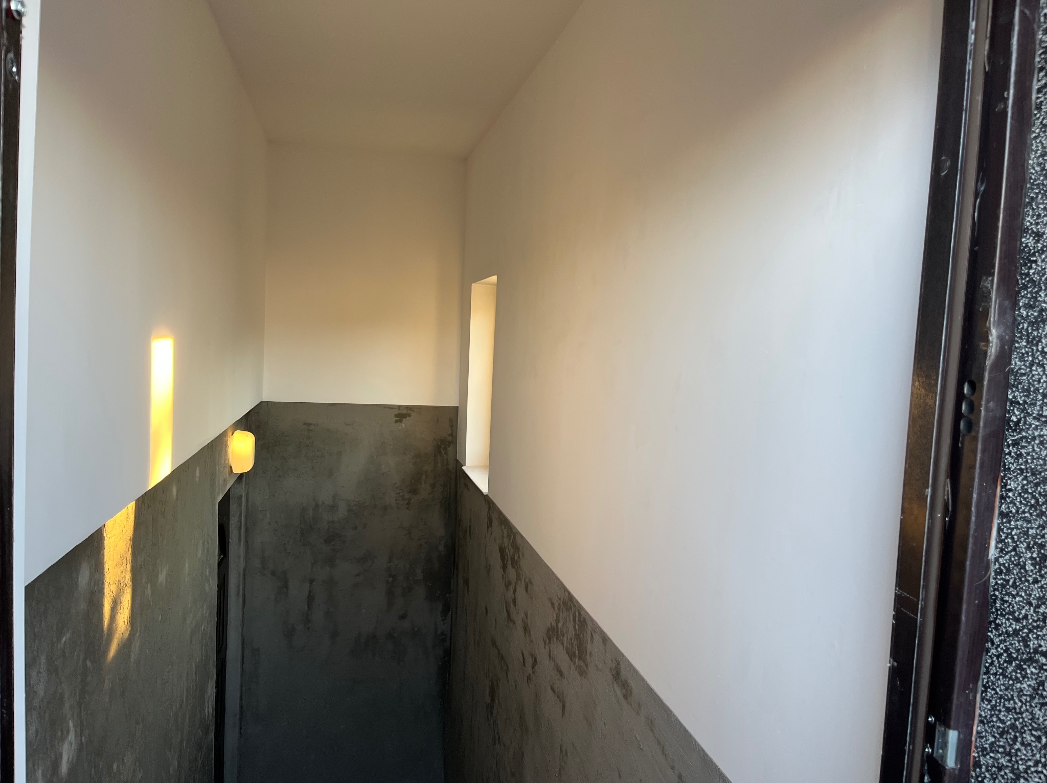 Wnętrze klatki schodowej po remoncie: białe ściany u góry, szare betonowe wykończenie na dole. Widoczne oświetlenie i fragment okna. Minimalistyczny design.