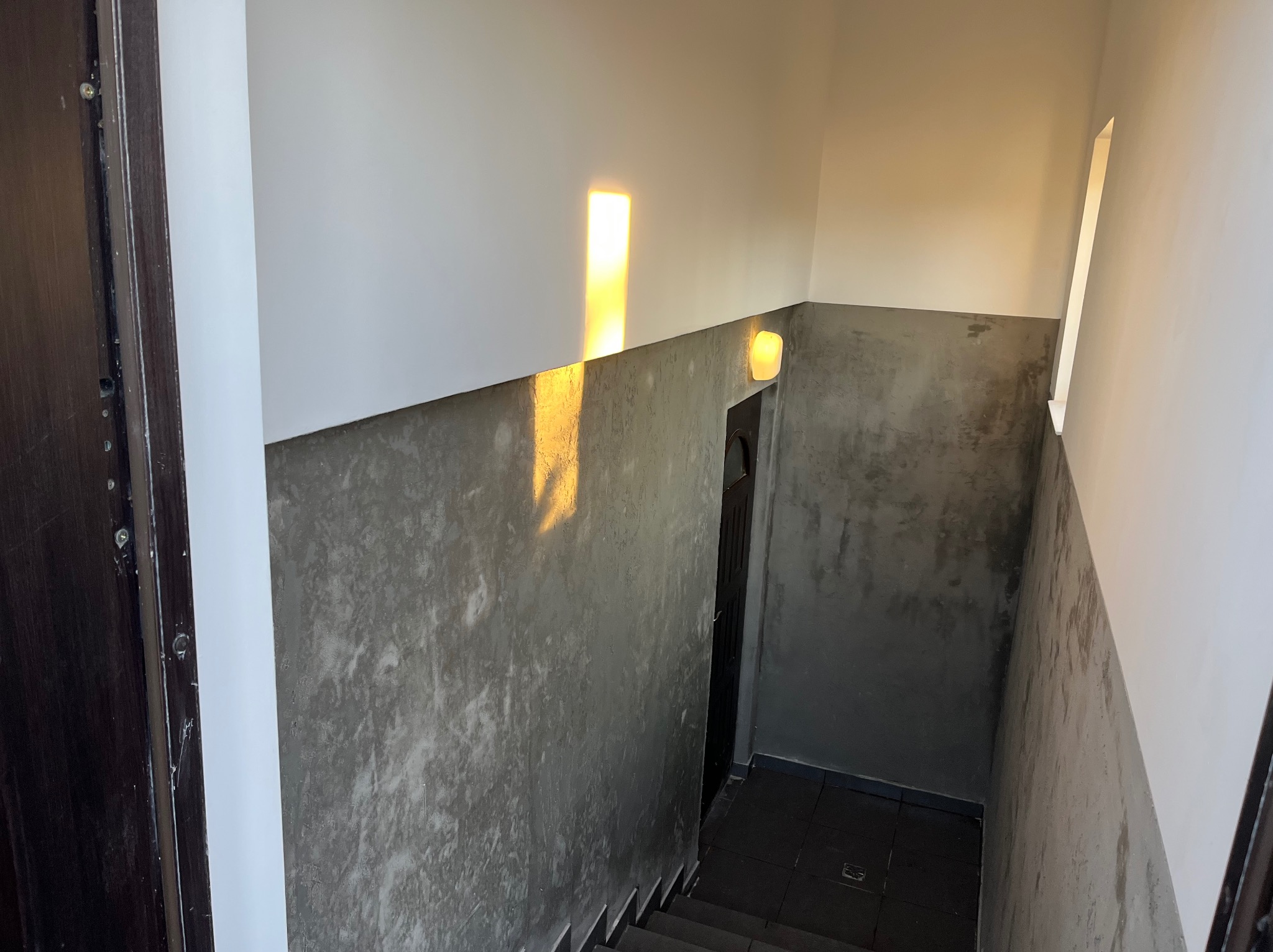 Klatka schodowa po remoncie: białe ściany u góry, szpachlowane na szaro do połowy wysokości. Wpadające światło słoneczne tworzy unikalny efekt wizualny.