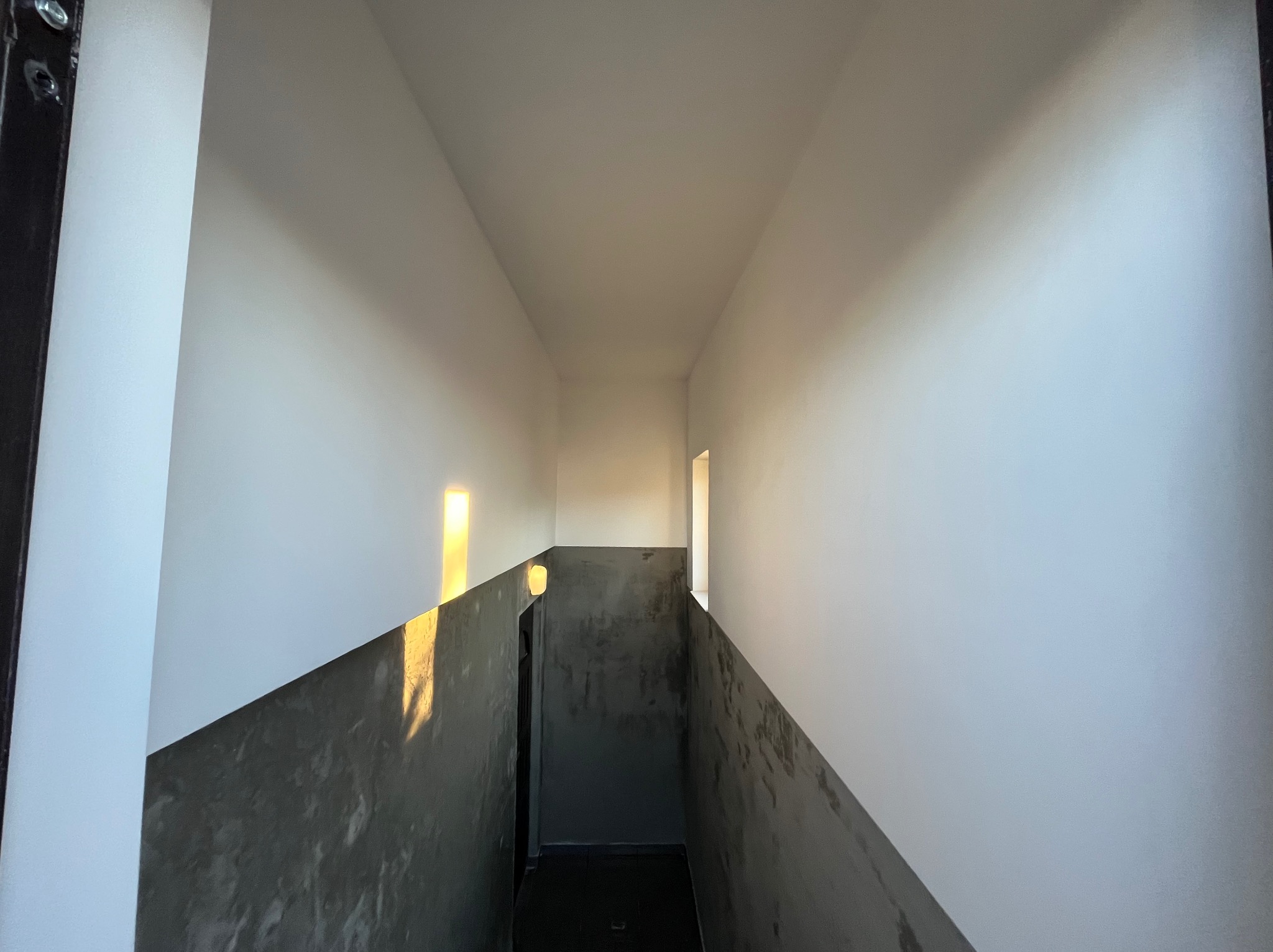 Wnętrze korytarza po szpachlowaniu i malowaniu ścian na biało, z fragmentem ściany pokrytej szarą, surową fakturą. Widoczne światło wpadające przez okno.