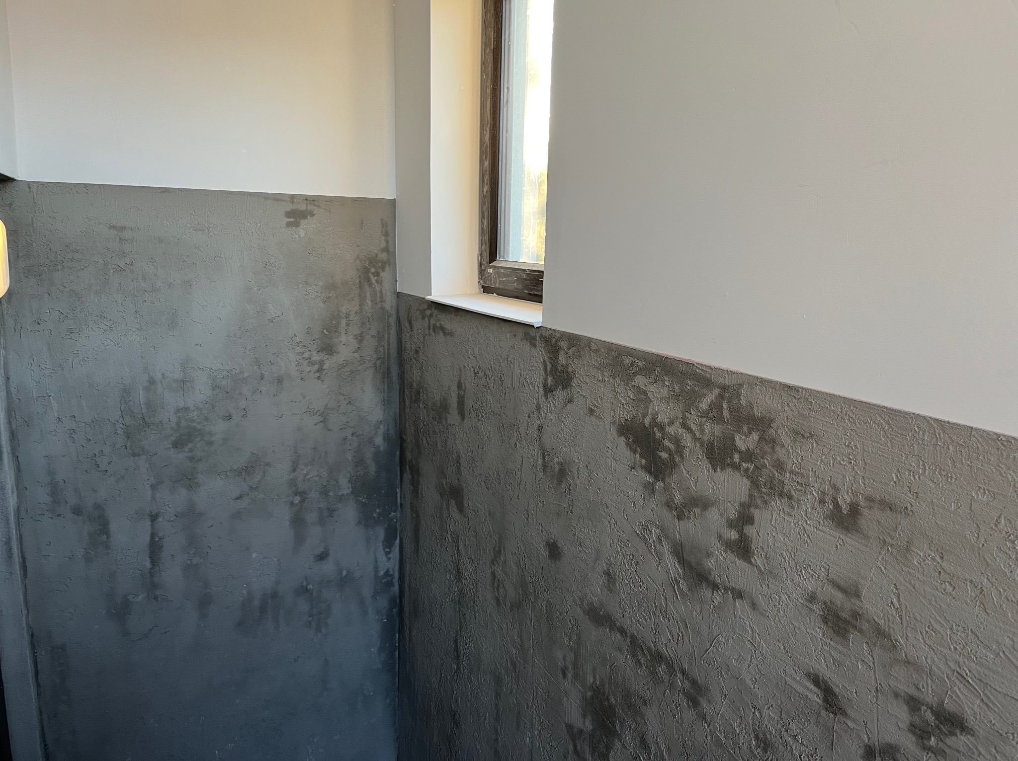 Ściana z efektem dekoracyjnym w odcieniach szarości, podzielona poziomą linią na dwie strefy kolorystyczne. W rogu widoczne okno z białym parapetem.