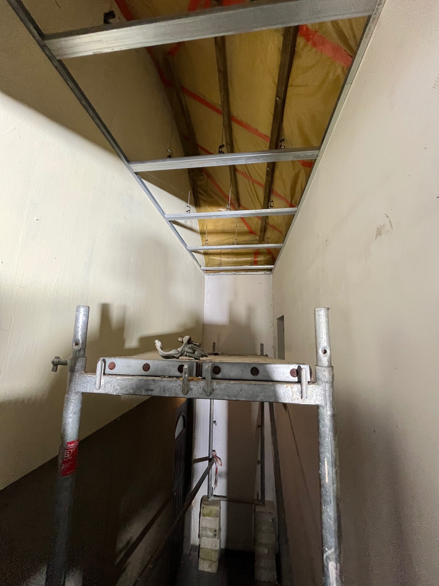 Montaż sufitu podwieszanego z płyt karton-gips, widok z rusztowania. Metalowa konstrukcja, izolacja z wełny mineralnej. Ściany w kolorze kremowym.