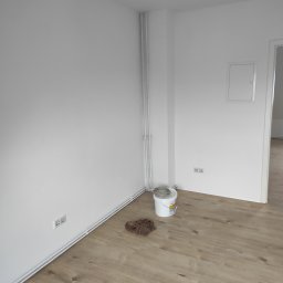 Lukasz Cichocki - Świeżo pomalowane na biało ściany w rogu pokoju z panelami podłogowymi w Rajczy. Widoczne puszka z farbą, grzejnik i gniazdka elektryczne.