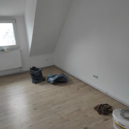 Lukasz Cichocki - Pomalowane na biało poddasze ze skosami, z workami na śmieci, grzejnikiem, puszką farby i szmatą na podłodze. Widok na okno i białe ściany.