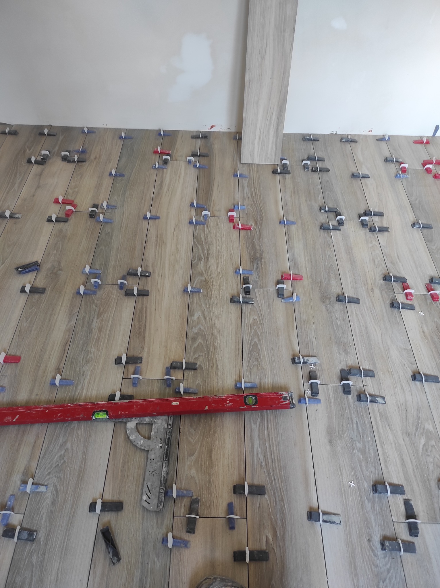 Ułożone płytki podłogowe imitujące drewno, z użyciem poziomicy i klipsów do wyrównywania, jedna płytka ustawiona pionowo przy ścianie.