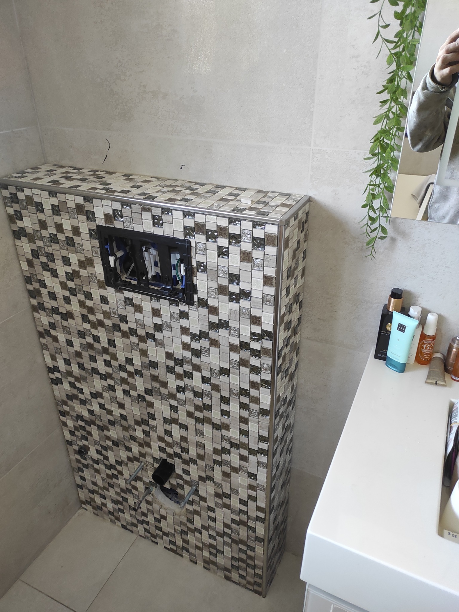 Obudowa stelaża WC z mozaiki w łazience, widoczne przyłącza. Umywalka z kosmetykami obok, lustro z zieloną girlandą w tle. Detal wykończenia.