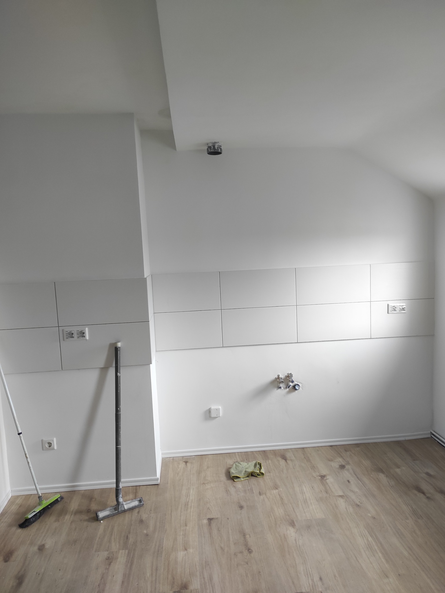 Biała kuchnia w trakcie wykończenia: płytki na ścianie, surowe przyłącza, drewnopodobna podłoga, narzędzia i ściereczka na podłodze. Minimalistyczny styl.