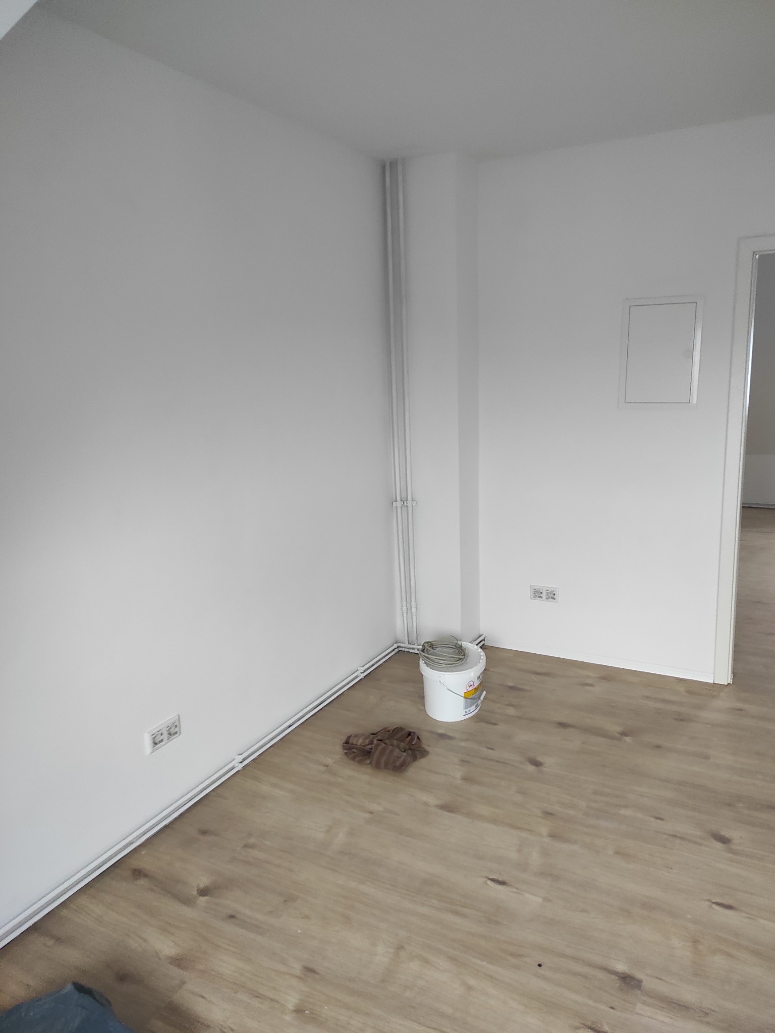Świeżo pomalowane na biało ściany w rogu pokoju z panelami podłogowymi w Rajczy. Widoczne puszka z farbą, grzejnik i gniazdka elektryczne.