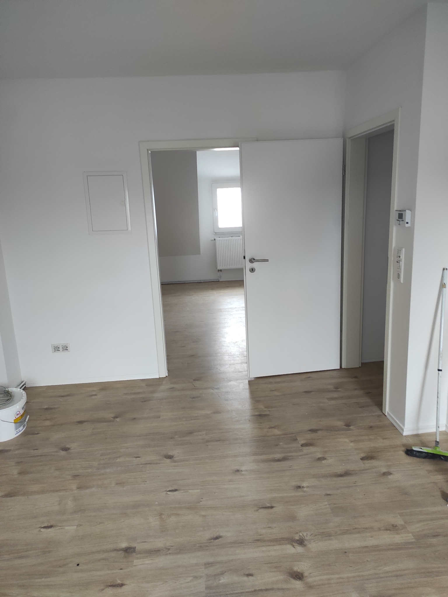 Jasne wnętrze po remoncie: drewnopodobna podłoga, białe ściany i drzwi. Widoczna puszka farby i sprzęt do sprzątania sugerują świeże wykończenie.