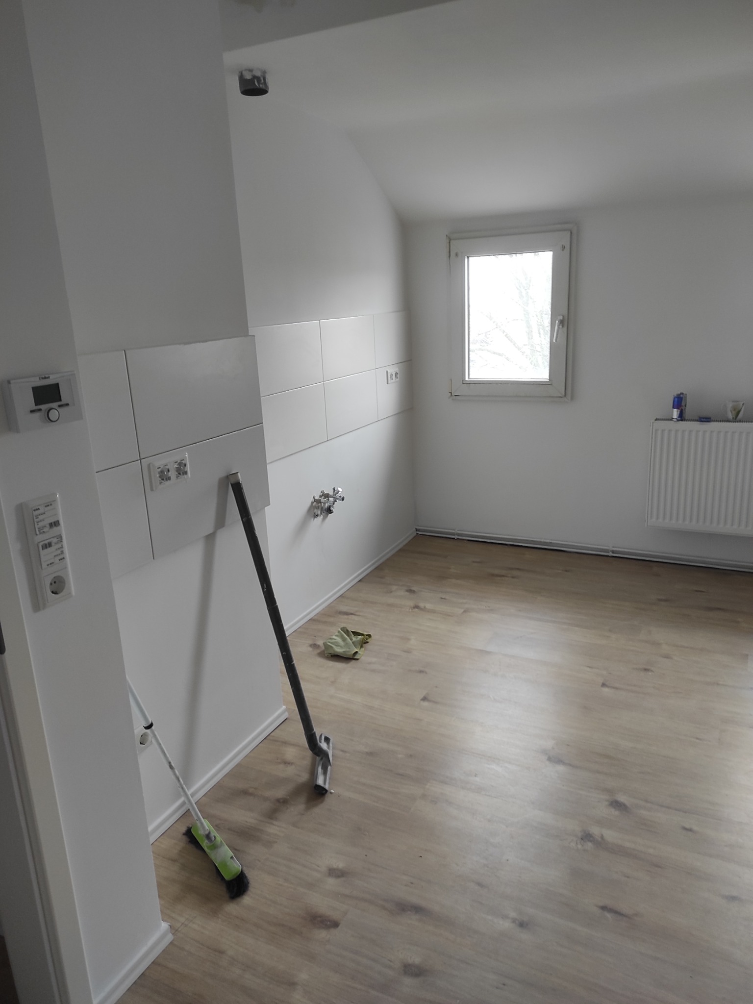 Biała kuchnia po remoncie z drewnianą podłogą, widoczne narzędzia i świeżo położone płytki. Minimalistyczny styl, jasne wnętrze z oknem.