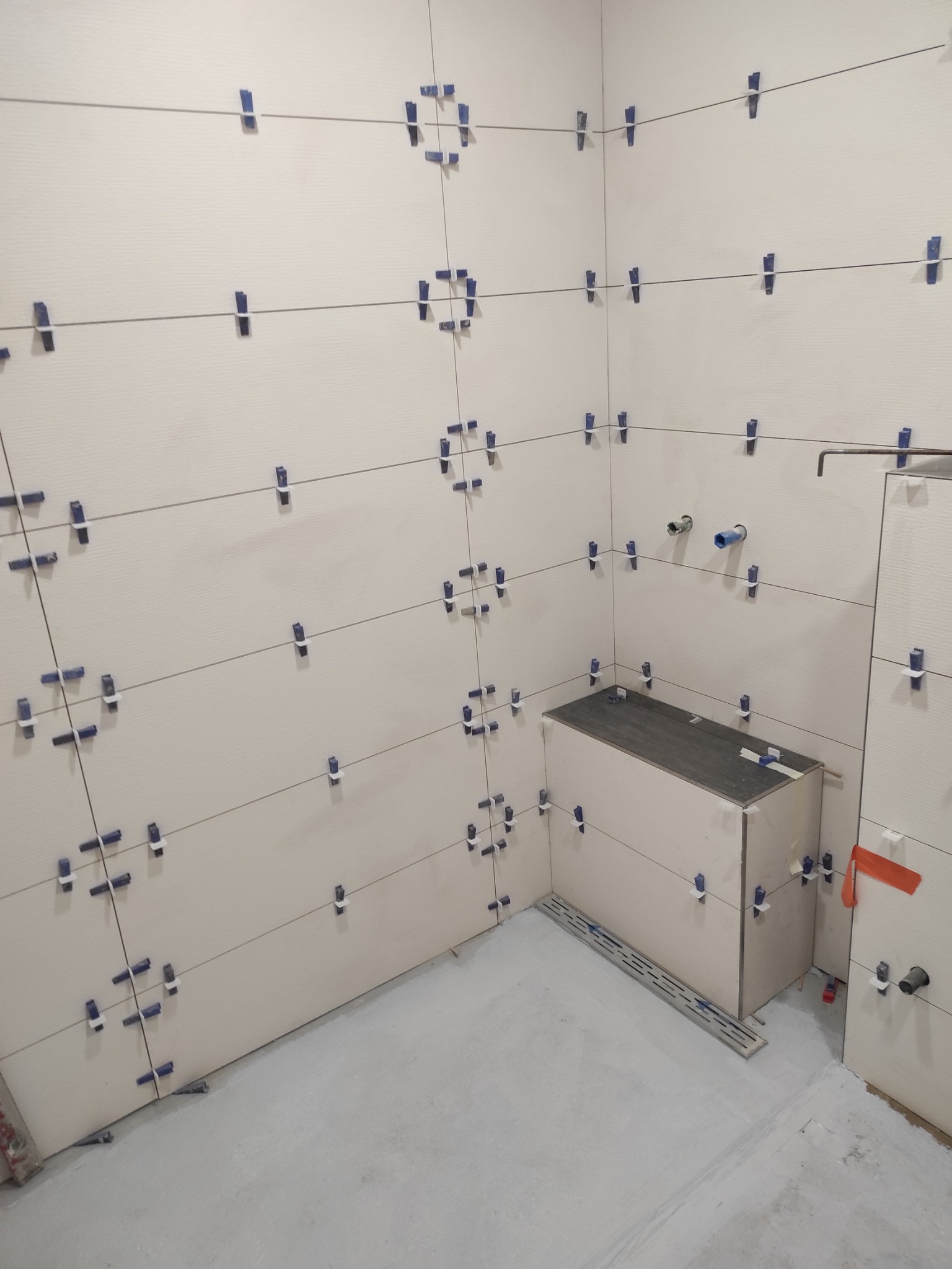 Ściany w trakcie układania płytek, z użyciem systemu poziomowania płytek. Widoczne niebieskie klipsy i sznurki. Fragment zabudowy z płytek. Szare fugi.