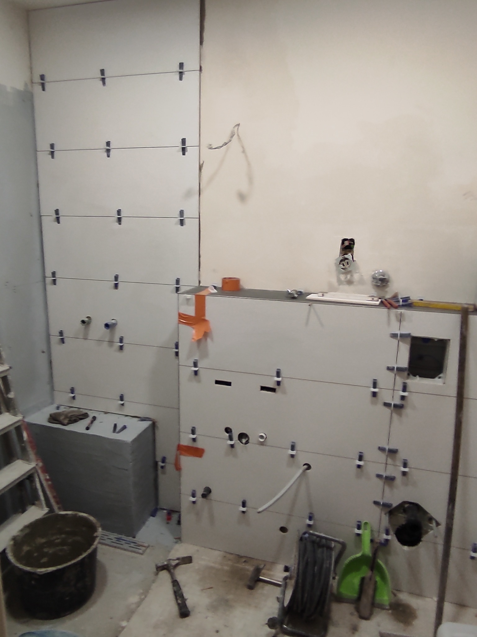 Proces układania białych płytek na ścianie, widoczne krzyżyki dystansowe, poziomica, narzędzia glazurnicze i przygotowane otwory na instalacje. Częściowo ukończona ściana.