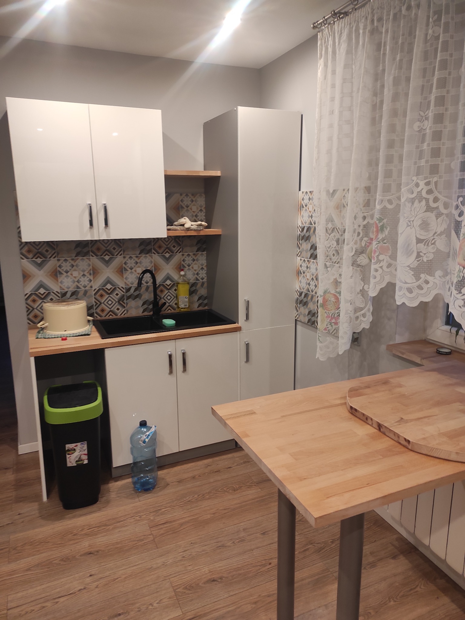 Jasna kuchnia z białymi szafkami, czarnym zlewem i wzorzystymi płytkami nad blatem. Drewniany stół z okrągłą nakładką i wzorzysta firanka w oknie.