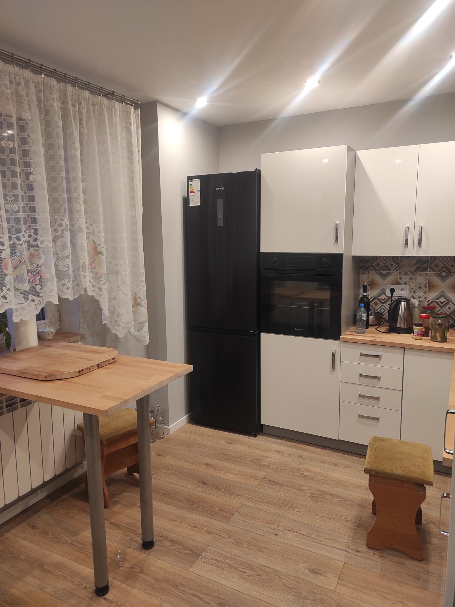 Nowoczesna kuchnia z czarną lodówką Gorenje, białymi szafkami i drewnianym blatem. Uwagę przyciąga wzorzysta ściana nad blatem i drewniana podłoga.