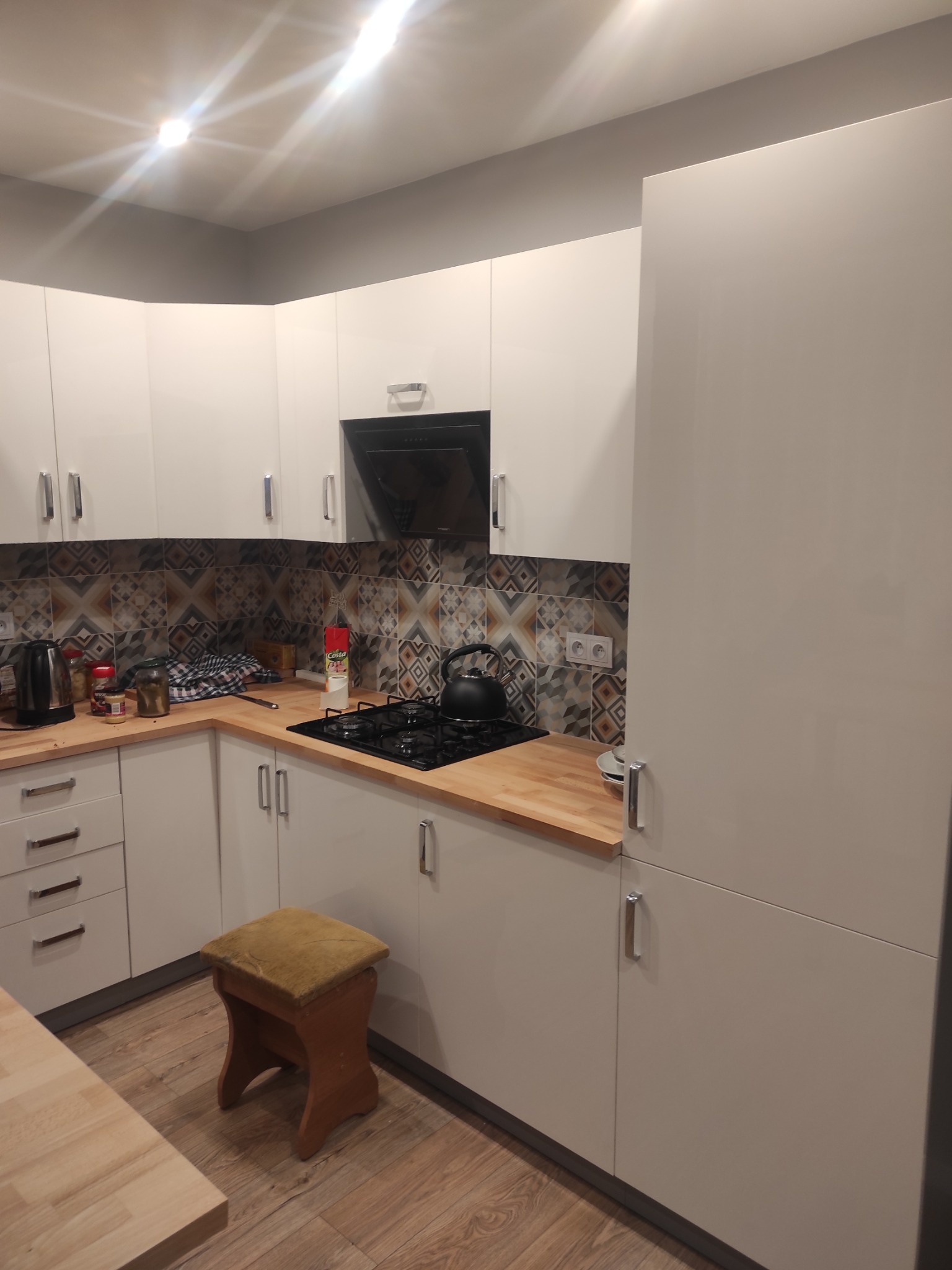 Jasna kuchnia z drewnianym blatem i wzorzystą mozaiką nad płytą grzewczą. Widoczny fragment stołu i stołek.