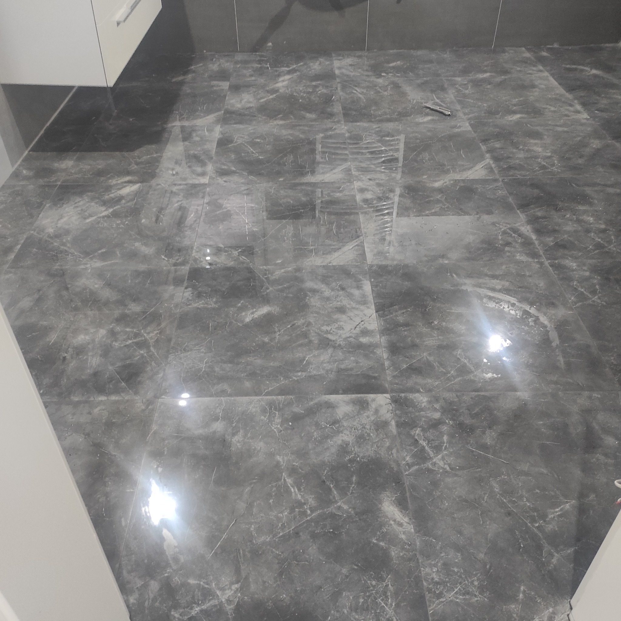 Błyszcząca, ciemnoszara podłoga z płytek imitujących marmur w kuchni. Widoczny fragment białej szafki i nóż na podłodze. Czyste, wykończone wnętrze.
