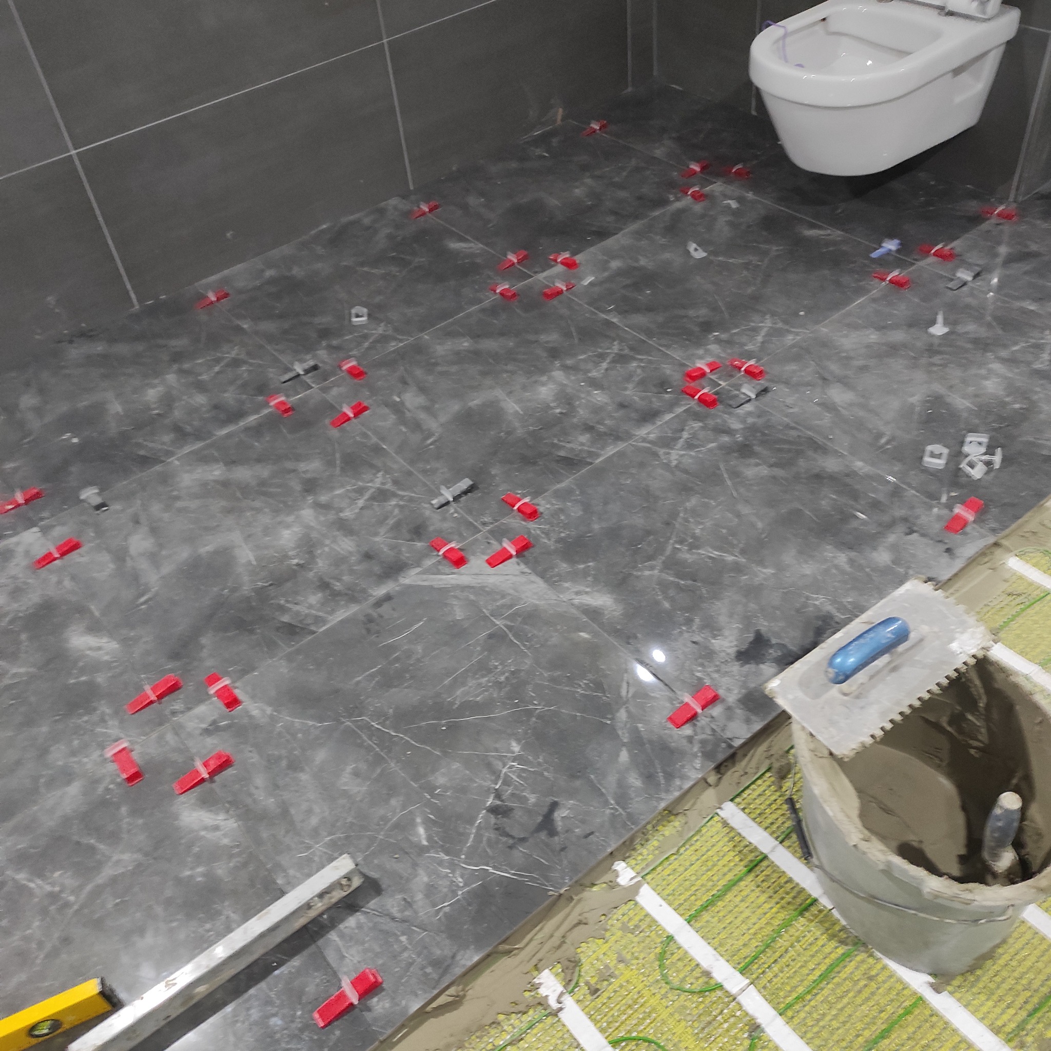 Etap układania ciemnych płytek podłogowych z marmurowym wzorem w łazience, widoczne poziomica, wiadro z klejem i system poziomowania płytek.