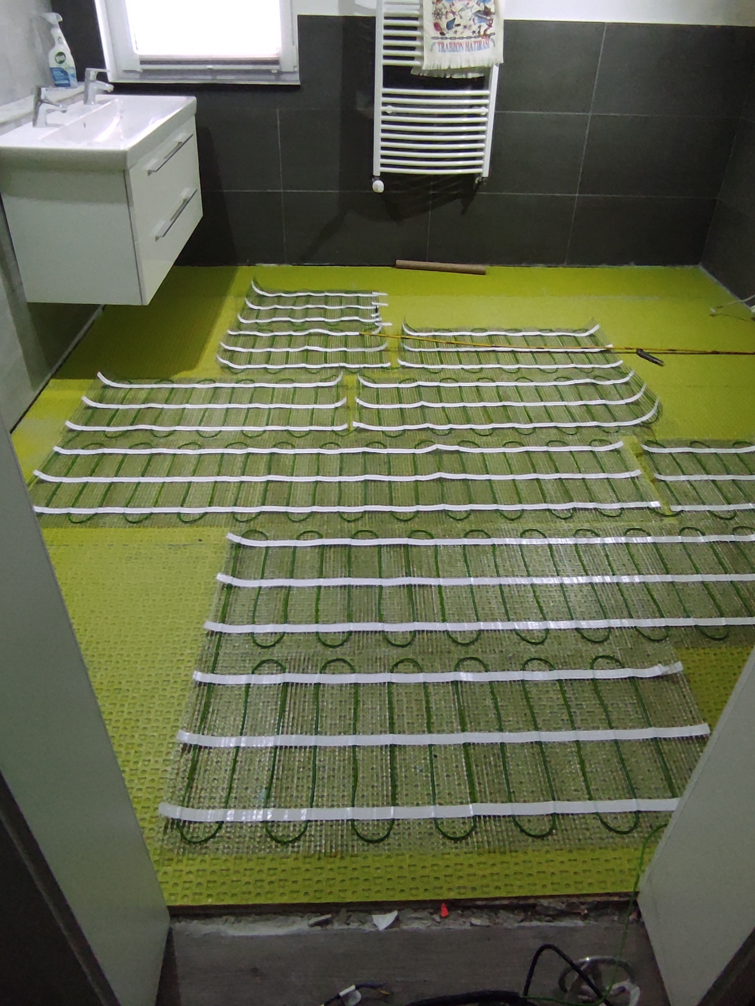 Rozłożona mata grzewcza na podłodze w łazience, przygotowana do wylewki. Widoczne elementy instalacji, płytki na ścianie i umywalka.